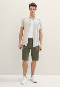 Camisa de botones de manga corta color beige claro, camiseta blanca debajo, pantalones cortos cargo verdes con bolsillos laterales y zapatillas blancas.