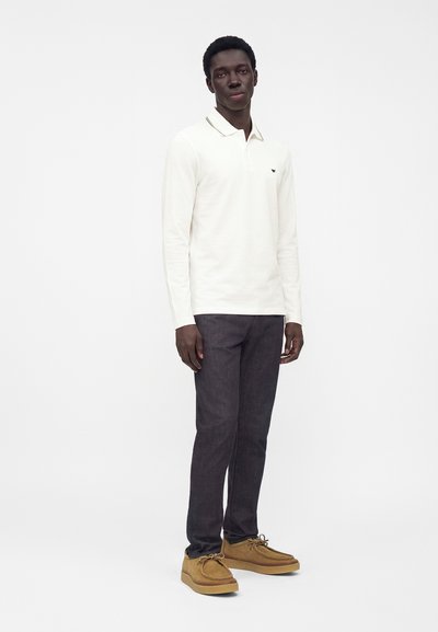 Homme debout portant un polo blanc à manches longues, un jean foncé et des chaussures à lacets couleur beige sur un fond blanc uni.