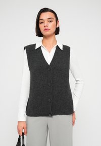 Γυναίκα με κοντά σκούρα μαλλιά που φοράει λευκό πουκάμισο με γιακά, σκούρο γκρι πλεκτό γιλέκο με κουμπιά, ανοιχτό γκρι παντελόνι και κρατάει μια μαύρη τσάντα χειρός.