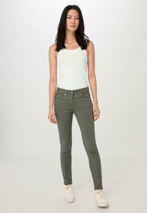 Olivgrüne Slim-Fit-Jeans mit Fronttaschen, einem Fünf-Taschen-Design und einer subtilen Textur, kombiniert mit einem weißen Tanktop und hellen Turnschuhen.