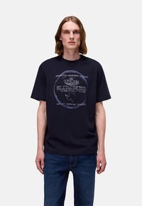 Marineblaues T-Shirt mit einem grafischen Aufdruck aus Text und Globus-Design. Kurze Ärmel und Rundhalsausschnitt. Kombiniert mit blauen Denim-Jeans.
