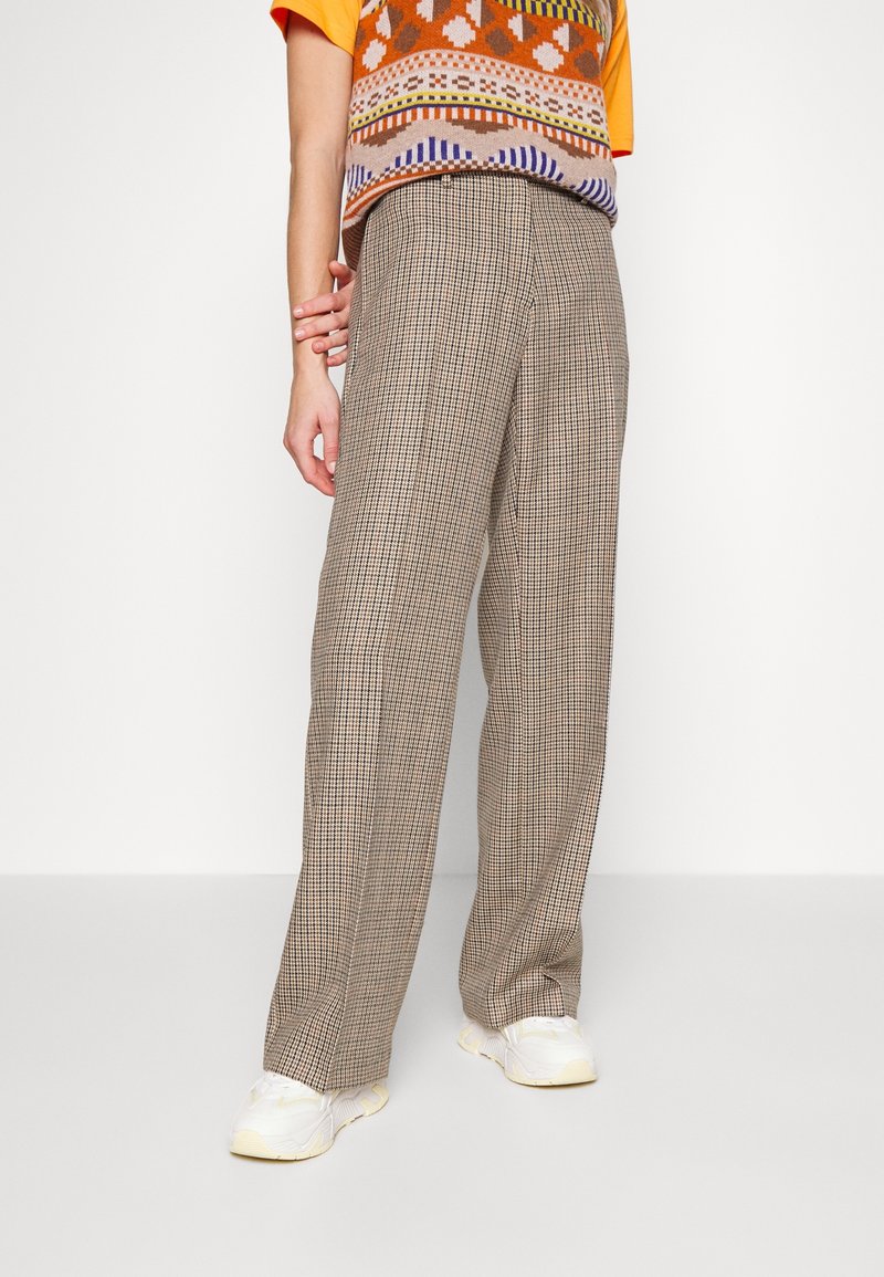 Personne portant un pantalon large à carreaux beige, des baskets blanches, un gilet à motif orange et une chemise jaune à manches courtes.