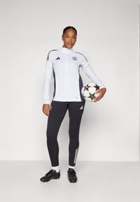 Ensemble de survêtement Adidas en blanc avec des accents violets, comprenant un col montant et des leggings noirs avec des rayures blanches, accompagné d'un ballon de football.