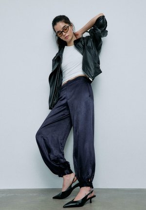 Žena u crnoj kožnoj jakni, bijelom kratkom topu, tamnim širim hlačama, crnim šiljastim potpeticama i obojenim naočalama, pozira naslonjena na sivu zid.