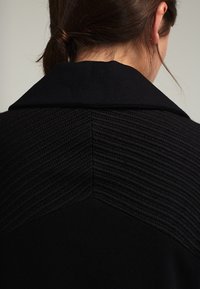Vue de dos d'une personne aux cheveux bruns attachés en un chignon bas, portant un manteau noir texturé avec un large col.