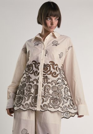 Kobieta ubrana w kremową koszulę na guziki i spodnie z brązową kwiatową koronkową aplikacją i falbankowym wykończeniem, patrząca na bok.