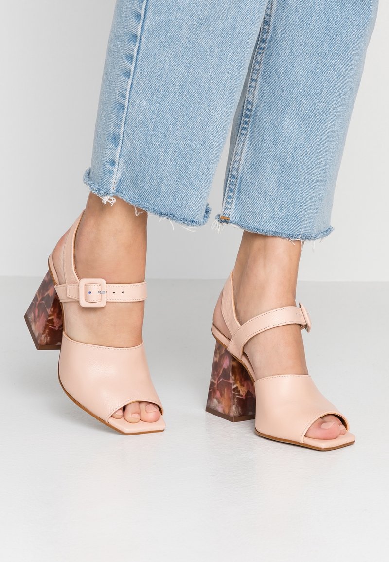 Nohy v světle růžových otevřených podpatcích s mramorovými blokovými podpatky a pružnými pásky kolem kotníků, doplněné o zkrácené světle modré potrhané džíny.