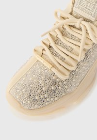 Zapatilla beige de Steve Madden con cordones gruesos, adornada con pequeñas perlas y piedras brillantes plateadas en la parte superior y la lengüeta.