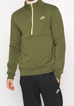 Mand iført olivengrøn Nike quarter-zip sweatshirt og sorte Nike sweatpants med logoer på bryst og lår.