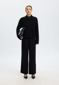 Juodas pūkuotas striukė su užtrauktuku ir aukštu apykakliu, dvi priekinės kišenės, derinama su plačiomis juodomis kelnėmis ir smailiais juodais aukštakulniais, rankoje laikoma juoda rankinė.