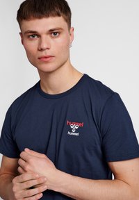 Mörkblå T-shirt gjord av bomull med rund hals. Har en röd och vit broderad logotyp på bröstet. Minimalistisk design, korta ärmar.