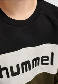 Černá, olivově zelená a bílá mikina se žebrovaným kulatým výstřihem. Obsahuje vodorovný bílý pruh s texturou černého písma "hummel."