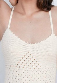 Vaalean kermanvärinen virkattu toppi, jossa on aaltoilevat reunat, timantinmuotoinen avokudontakuosi ja ohuet hihnat.