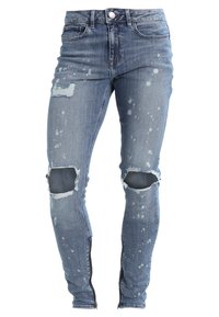 Jean slim-fit en denim bleu avec genoux usés, détails de taches de peinture blanche, bouton à l’avant, braguette zippée et fermetures éclair aux chevilles.