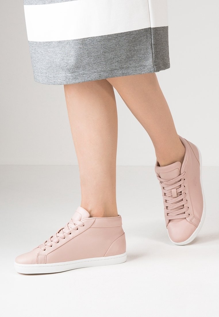 Jambes portant une jupe grise et blanche avec des baskets montantes rose clair, debout sur un sol blanc devant un fond clair.