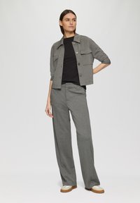 Grijze cropped jack met twee voorzakken, gedragen met een zwarte ribbeltop en losse grijze broek. Model draagt beige en bruine schoenen.