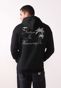 Sudadera con capucha de forro polar negro que presenta una capucha, puños acanalados y dobladillo. El diseño en la parte posterior incluye palmeras y el texto "Naturaleza en Armonía" en blanco.