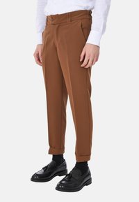 Pantaloni marroni su misura con un tessuto morbido, caratterizzati da una vita con bottoni e risvolti, abbinati a scarpe e calzini neri.