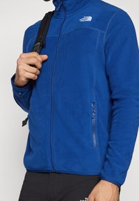 Man med blå fleecejacka med dragkedjefickor och svart ryggsäck hängande över axeln, beskuren vy.