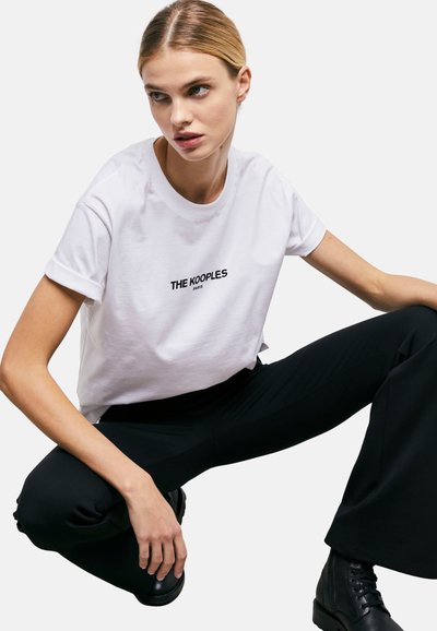 Balta kokvilnas T-kreklu ar sarullētām piedurknēm un melnu uzdrukātu tekstu "THE KOOPLES PARIS", kombinēta ar melniem biksēm un kurpēm ar �šņorēm virs potītes.