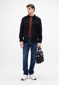 Giacca camicia in corduroy nero, maglione a coste color ruggine, jeans blu, sneaker bianche, mentre tiene una borsa nera con zip e manici.