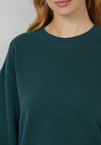 Femme portant un pull à col rond côtelé vert foncé, visible du cou à mi-poitrine, avec une peau claire et des cheveux blonds ondulés.