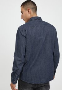 Camisa de mezclilla oscura con botones, color cuello, mangas largas y corte recto. Presenta costuras sutiles y botones en los puños.
