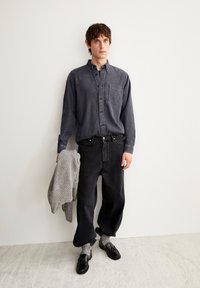 Camicia di denim nera con lavaggio precedente, caratterizzata da una tasca sul petto, abbinata a jeans neri larghi. Maglione grigio in maglia appoggiato sul braccio, scarpe nere indossate con calzini grigi.