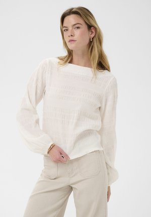 Jeune femme aux cheveux clairs portant un haut texturé blanc à manches longues et un pantalon beige, posant avec une main sur la hanche.