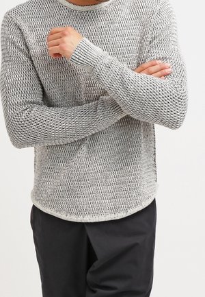 Mann verschränkt die Arme, trägt einen strukturierten hellgrauen Strickpullover und schwarze Hose, steht vor einem schlichten weißen Hintergrund.