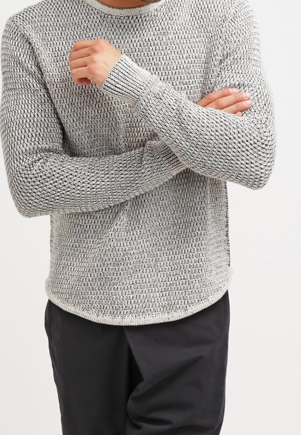 Homme les bras croisés, portant un pull en tricot gris clair texturé et un pantalon noir, debout devant un fond blanc uni.