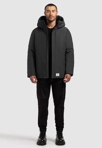 Black hooded parka jacket made of waterproof material, featuring a front zipper, side pockets, and a clean design. Paired with black cargo pants.