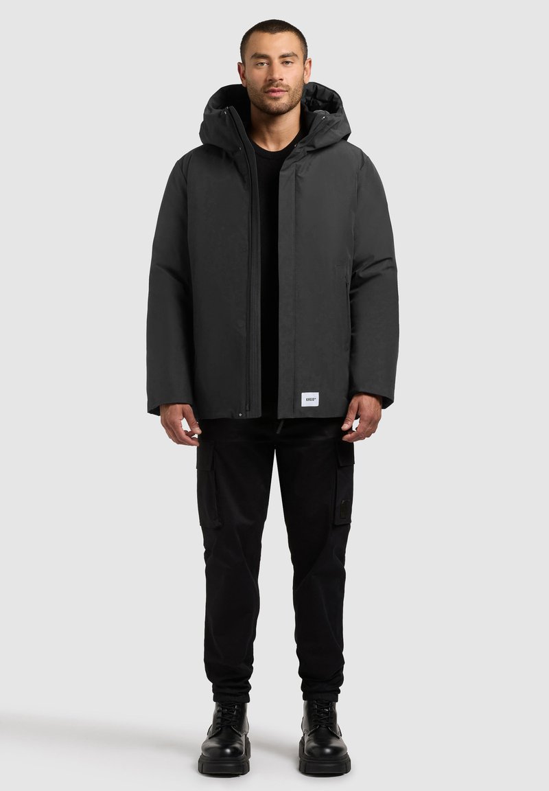 Black hooded parka jacket made of waterproof material, featuring a front zipper, side pockets, and a clean design. Paired with black cargo pants.