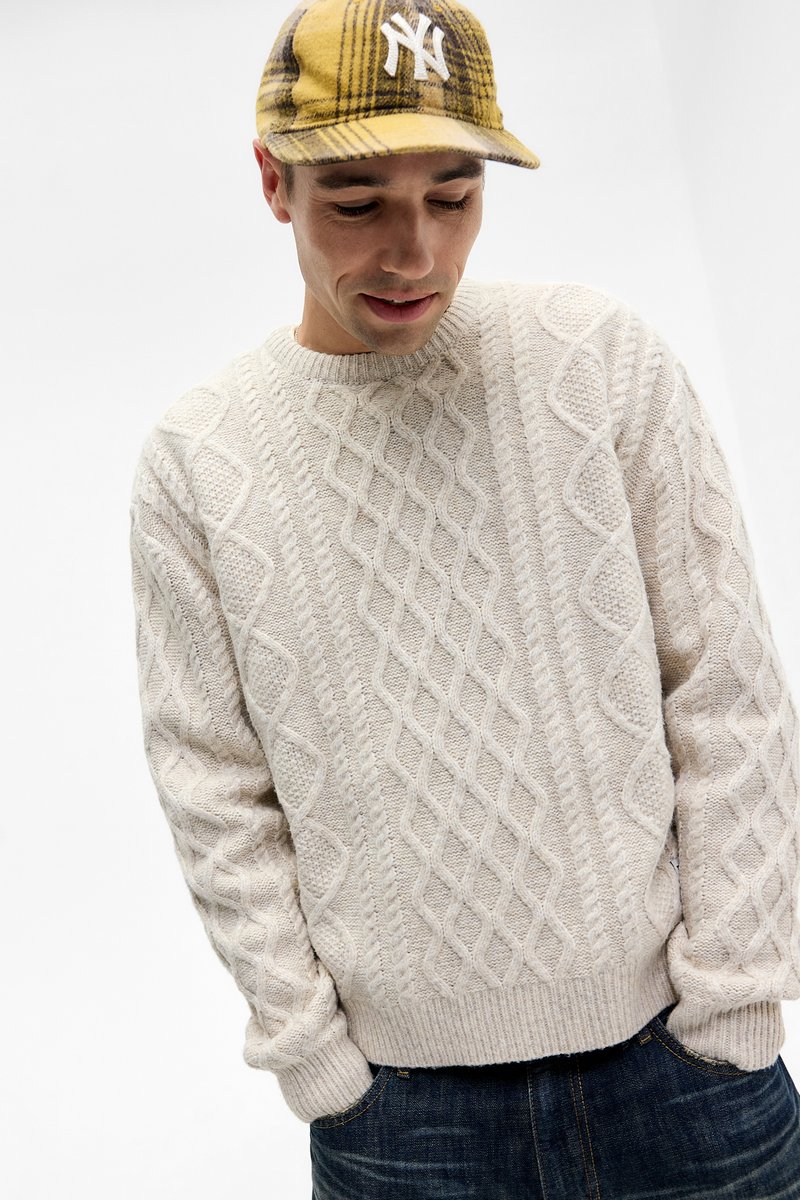 Cremefarvet kabelstrik sweater med rund halsudskæring og ribbede manchetter, stylet med mørkeblå jeans. Sweateren har strukturerede diamantmønstre.