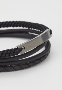 Bracelet en cuir tressé noir avec trois brins, un design tissé texturé et un accent de fermoir métallique lisse.