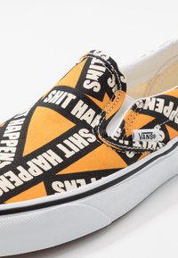 Baskets Vans à enfiler avec un motif orange et noir comportant un texte blanc en gras disant "SHIT HAPPENS" et une petite étiquette logo Vans sur le côté.