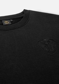 Zwarte sweatshirt met een geribde ronde hals en een zwarte, geborduurde logo op de borst. Het materiaal lijkt zacht en glad te zijn.