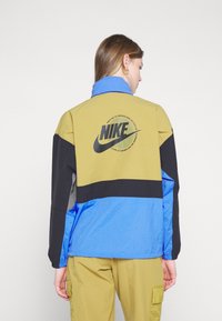 Nike-jakke med fargeblokker i olivengrønn, svart og blå. Baksiden har en stor svart logo i sirkulært design. Teksturert stoff, avslappet passform.
