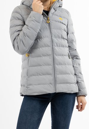 Person trägt eine hellgraue, gesteppte Daunenjacke mit gelben Reißverschlussanhängern und blauer Jeans und richtet den Jackenkragen mit einer Hand.