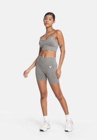 Šedý športový podprsenka a súprava vysokých šortiek z mäkkej, textúrovanej tkaniny; obsahuje minimálne logo; modelka je v pozícii s uvoľnenými rukami.