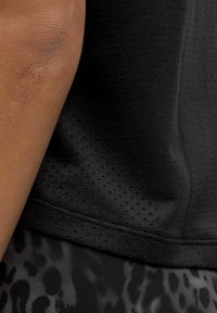 Canottiera sportiva nera in un tessuto testurizzato e traspirante con piccole perforazioni; un tessuto a motivi sottostante è parzialmente visibile.