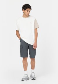 T-shirt blanc et beige à manches courtes avec un logo, associé à un short cargo noir. Les accessoires comprennent des chaussettes blanches et des chaussures de sport grises.
