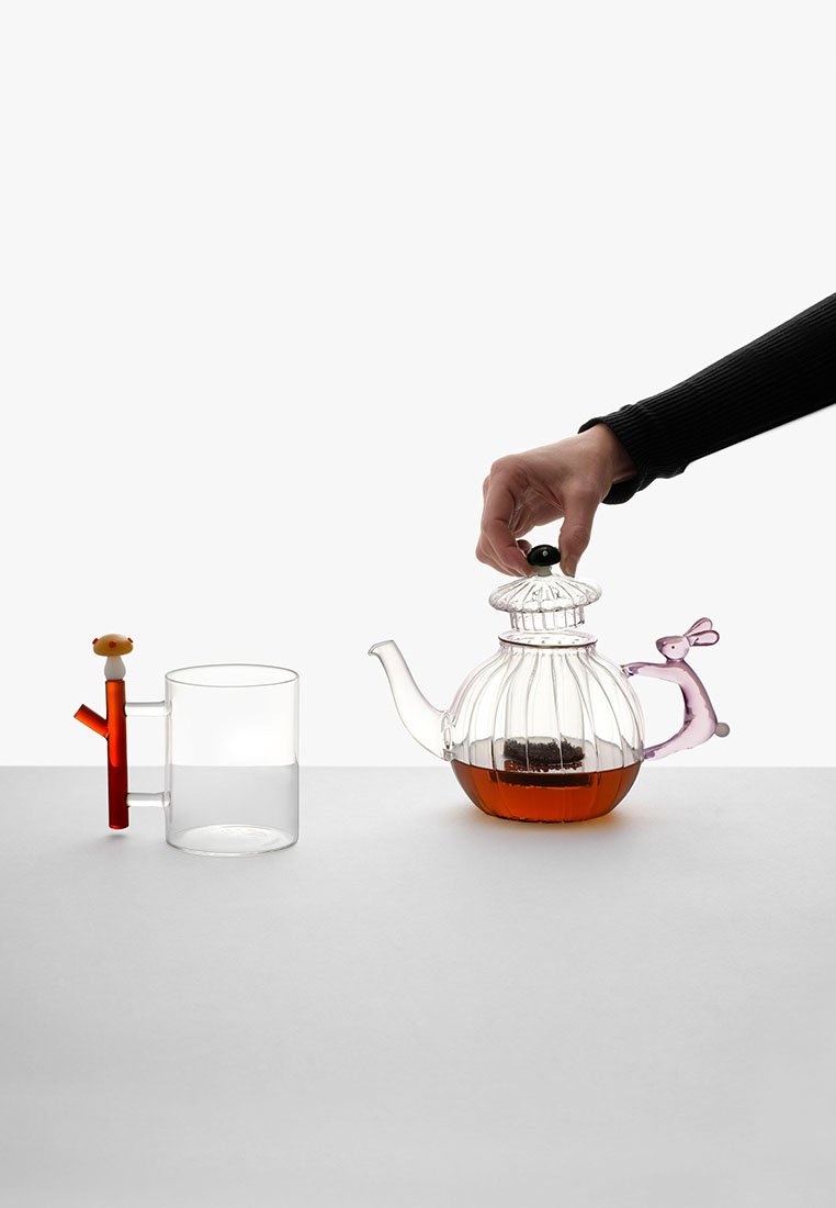 Przezroczysty szklany czajnik o żłobkowanej powierzchni, fioletowy uchwyt przypominający królika; obok przezroczysta szklanka z czerwonym uchwytem przypominającym słomkę.