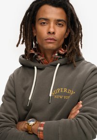 Grijze hoodie met verstelbare koorden en gele geborduurde tekst "SUPERDRY NEW YORK," gelaagd over een rood geruit shirt. Gouden horloge om de pols.
