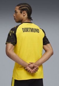 Persona con trenzas que lleva una camiseta deportiva amarilla y negra del Dortmund, con las manos entrelazadas detrás de la espalda, frente a un fondo neutro.