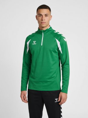 Ung mand iført grøn sportstrøje med lange ærmer med hvide accenter og sorte bukser, stående mod en ensartet lys baggrund.