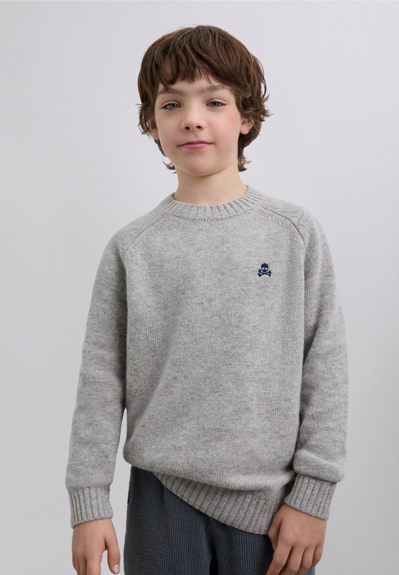 Suéter de punto gris claro con puños y dobladillo acanalados, con un pequeño emblema de calavera y huesos cruzados en azul marino en el pecho.