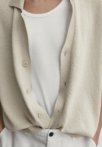 Beige gebreide cardigan met een losse fit, met knopen aan de voorkant over een witte geribde tanktop. Lichte textuur en ontspannen ontwerp.
