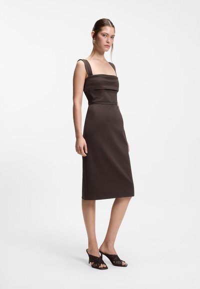 Femme portant une robe sans manches, brun foncé, longueur genou avec de larges bretelles, et des sandales à talons noires ouvertes aux orteils, debout devant un fond blanc.