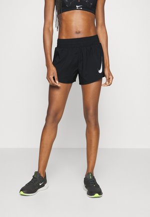 Woman wearing black Nike athletic shorts, black sports bra, and black running shoes with neon green accents, standing on a plain background.
